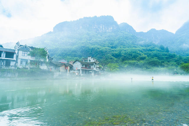 吉首市矮寨镇坪朗村，青山绿水环抱，河面薄雾缭绕，村民在石渡行走，苗寨宛如仙境