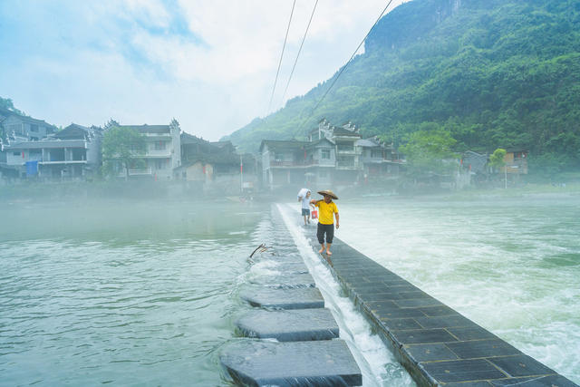 吉首市矮寨镇坪朗村，青山绿水环抱，河面薄雾缭绕，村民在石渡行走，苗寨宛如仙境
